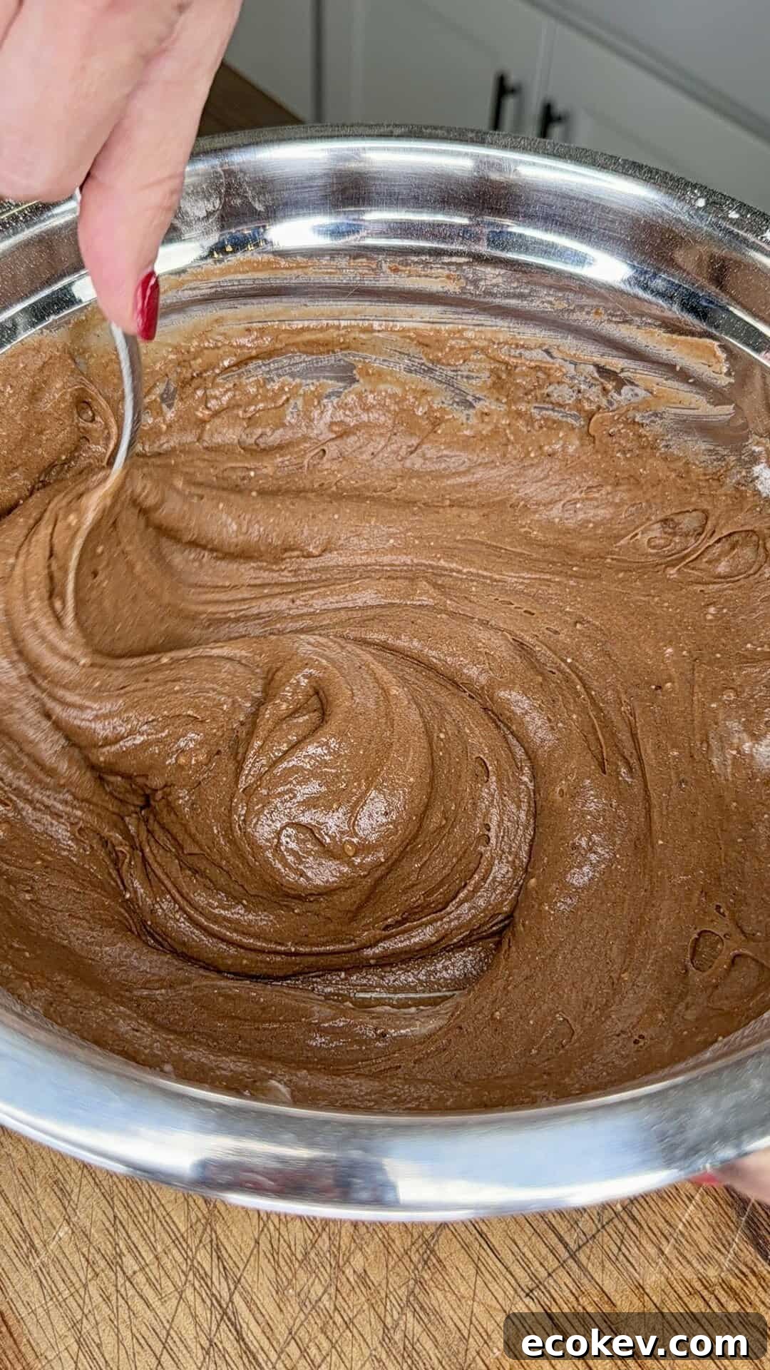 A hand with red nail polish stirs chocolate cake batter in a large metal mixing bowl using a spoon. The bowl is on a wooden surface in a kitchen.