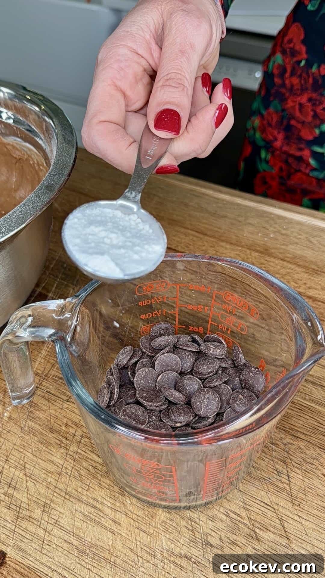 A hand with red nail polish holds a spoon of white powder above a measuring cup filled with chocolate chips on a wooden surface. A metal mixing bowl is partially visible to the left.