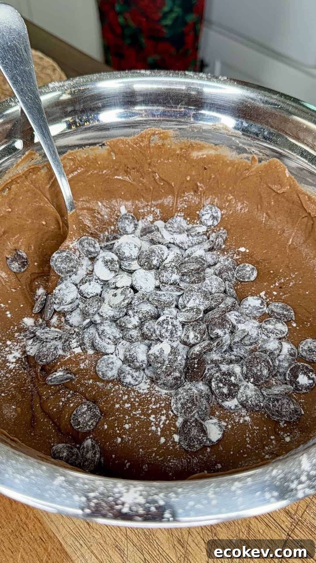 A metal bowl filled with chocolate cookie dough, topped with chocolate chips and a dusting of flour, sits on a wooden surface. A metal spoon is sticking out of the bowl.