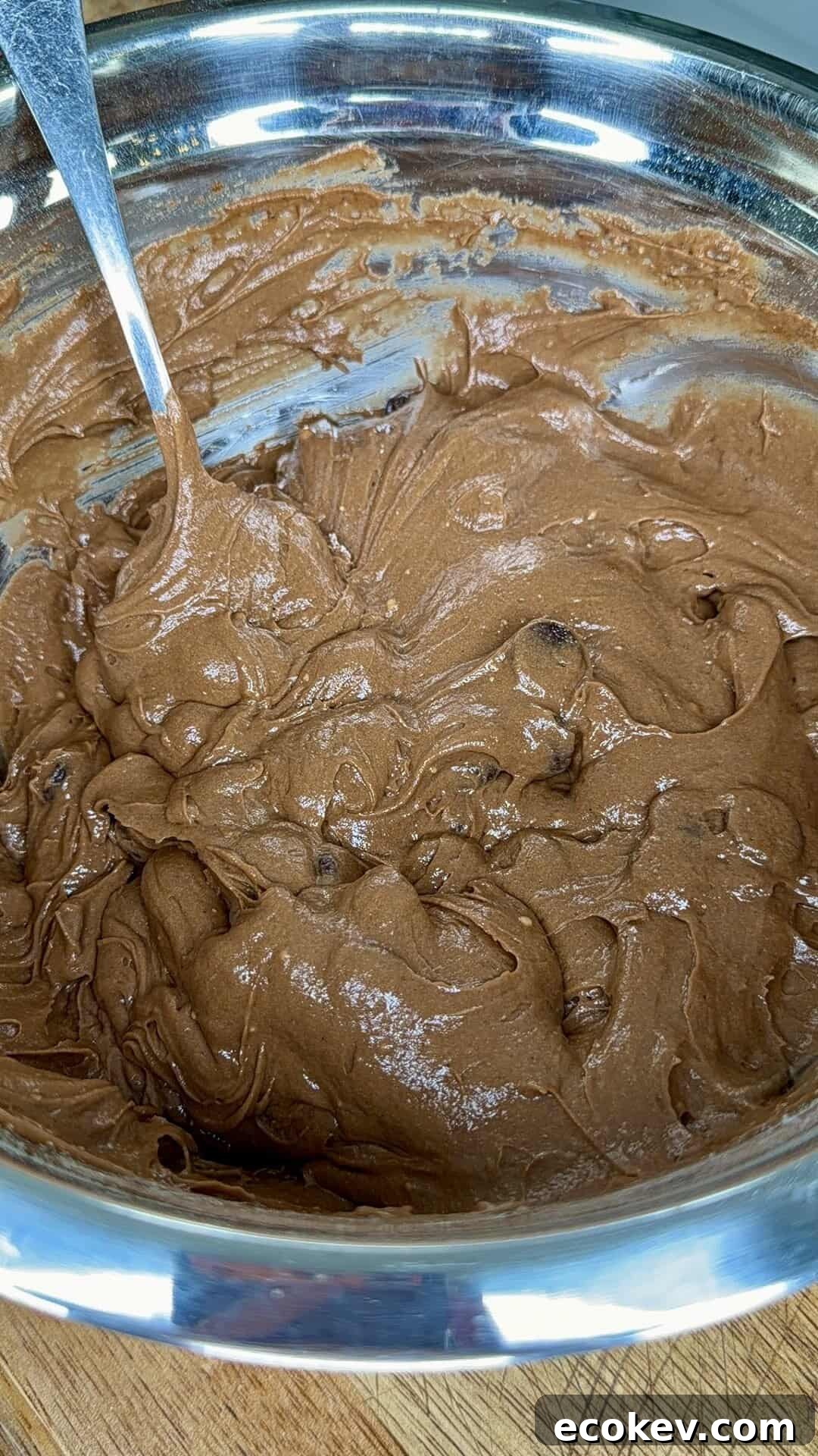 A metal bowl filled with thick, creamy chocolate batter being mixed with a metal spoon, sitting on a wooden surface.