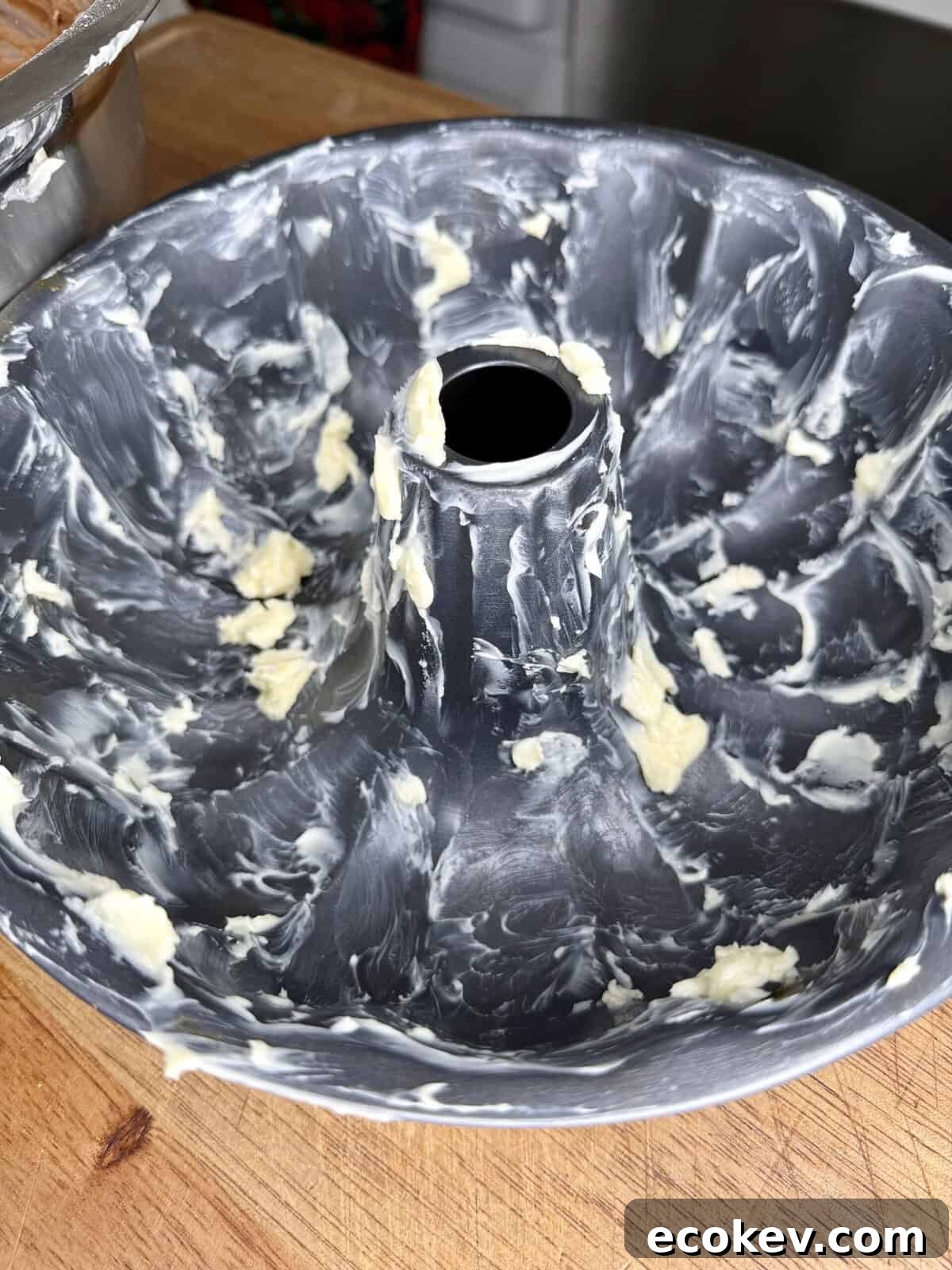 A close-up of a bundt cake pan coated with pats and smears of butter, sitting on a wooden surface, ready for cake batter.