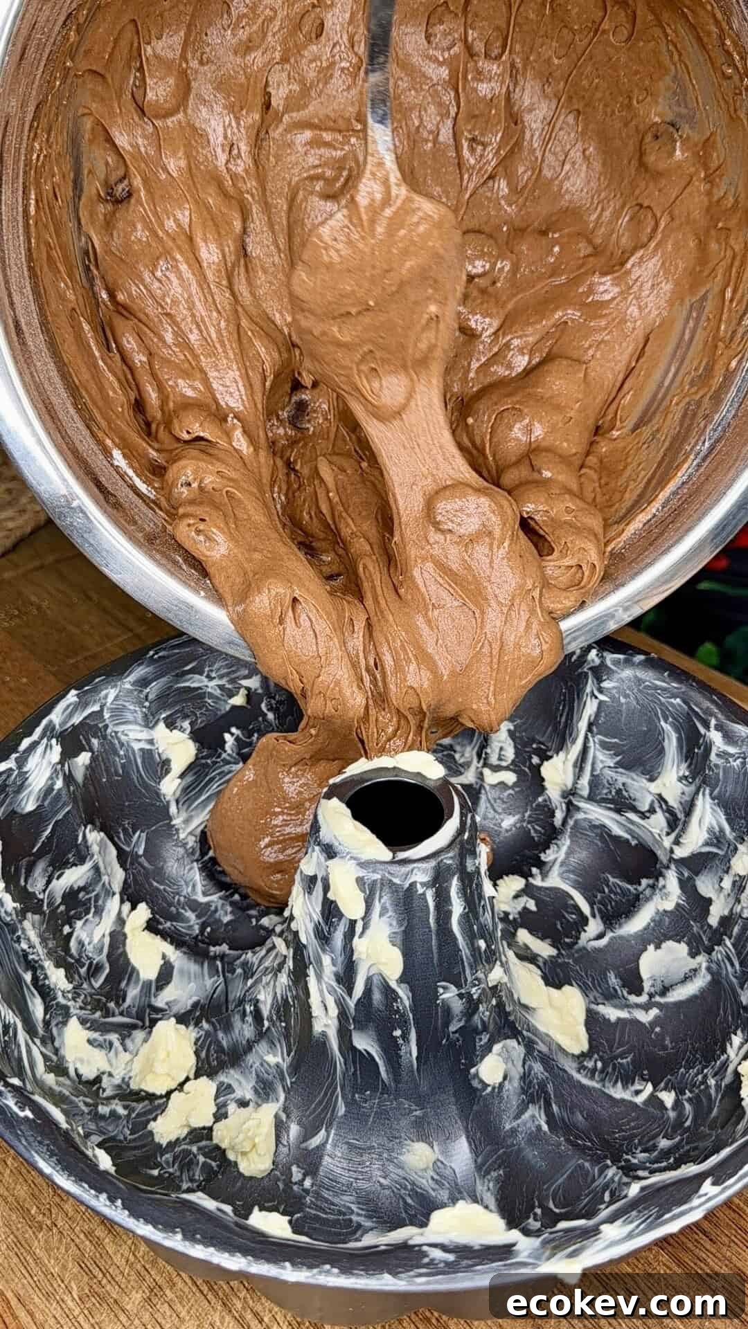 A thick chocolate cake batter is being poured from a metal bowl into a greased bundt pan, ready for baking.