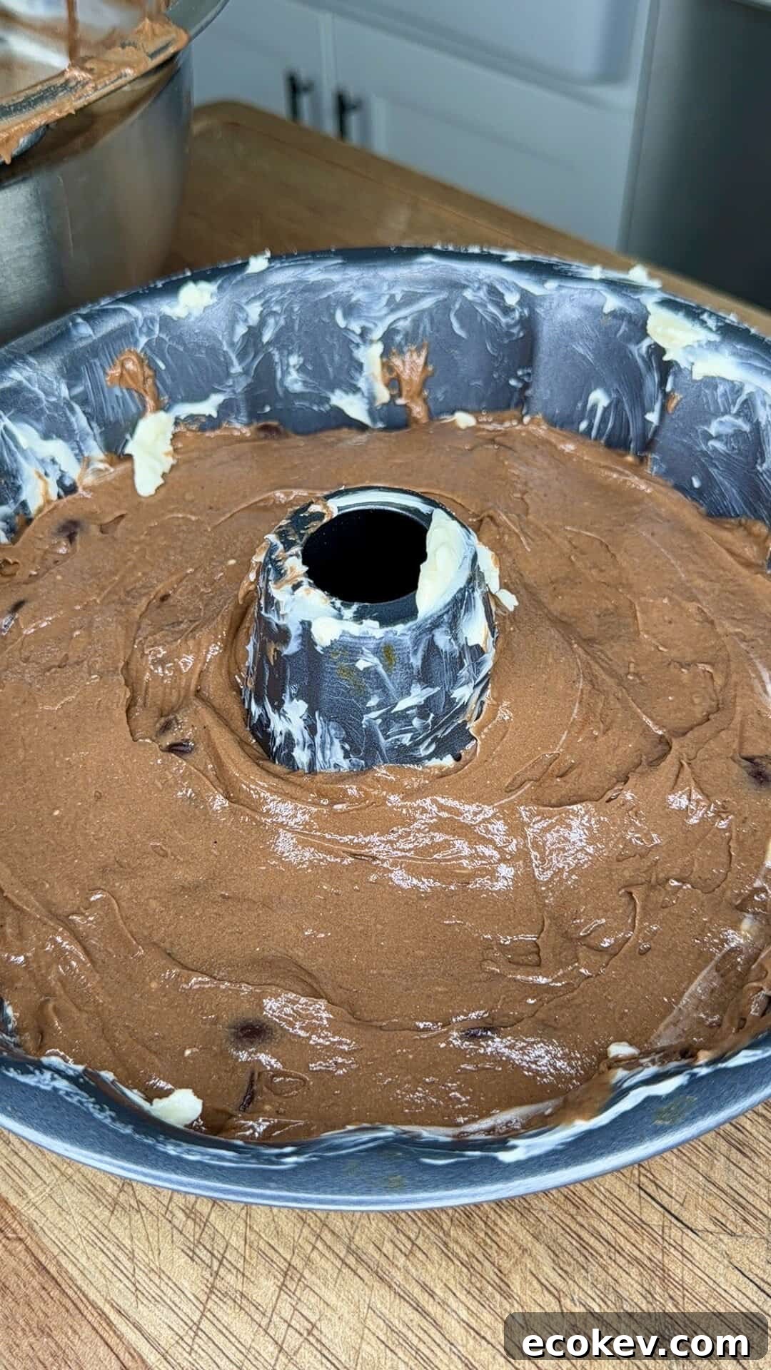 A bundt cake pan filled with chocolate cake batter sits on a wooden surface. The pan is greased with visible streaks of butter or shortening. In the background, a mixing bowl with more batter is partially visible.