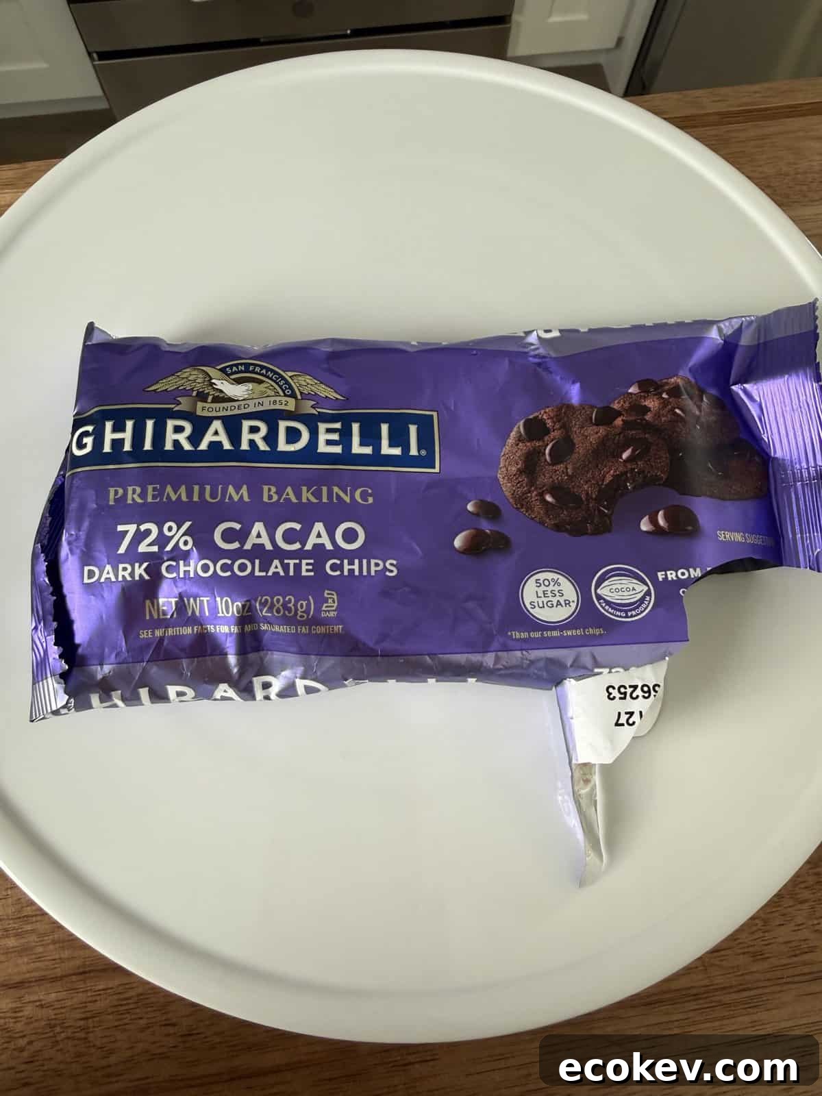 A partially opened bag of Ghirardelli 72% cacao dark chocolate chips sits on a white plate. The purple packaging displays images of chocolate chips and a cookie, with baking and cacao content details visible.