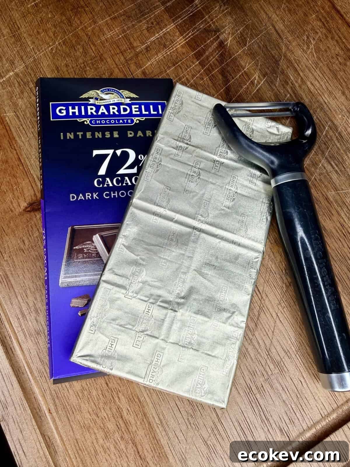 A Ghirardelli Intense Dark 72% Cacao chocolate bar, partly unwrapped, lies on a wooden surface next to a black-handled vegetable peeler.