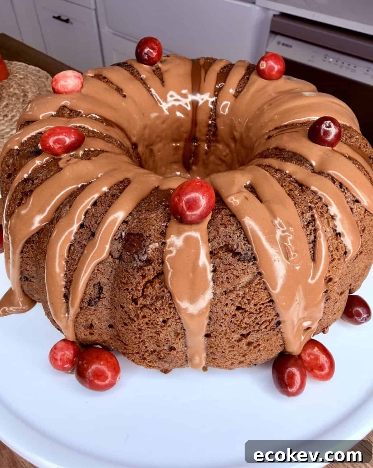 A bundt cake with light brown glaze drizzled on top, decorated with fresh red cherries, sits on a white plate in a kitchen setting.