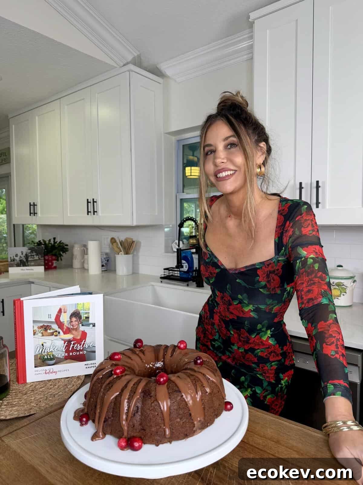 A smiling woman in a floral dress stands in a bright kitchen beside a chocolate bundt cake with cranberries and a cookbook on the counter. White cabinets and a window appear in the background.