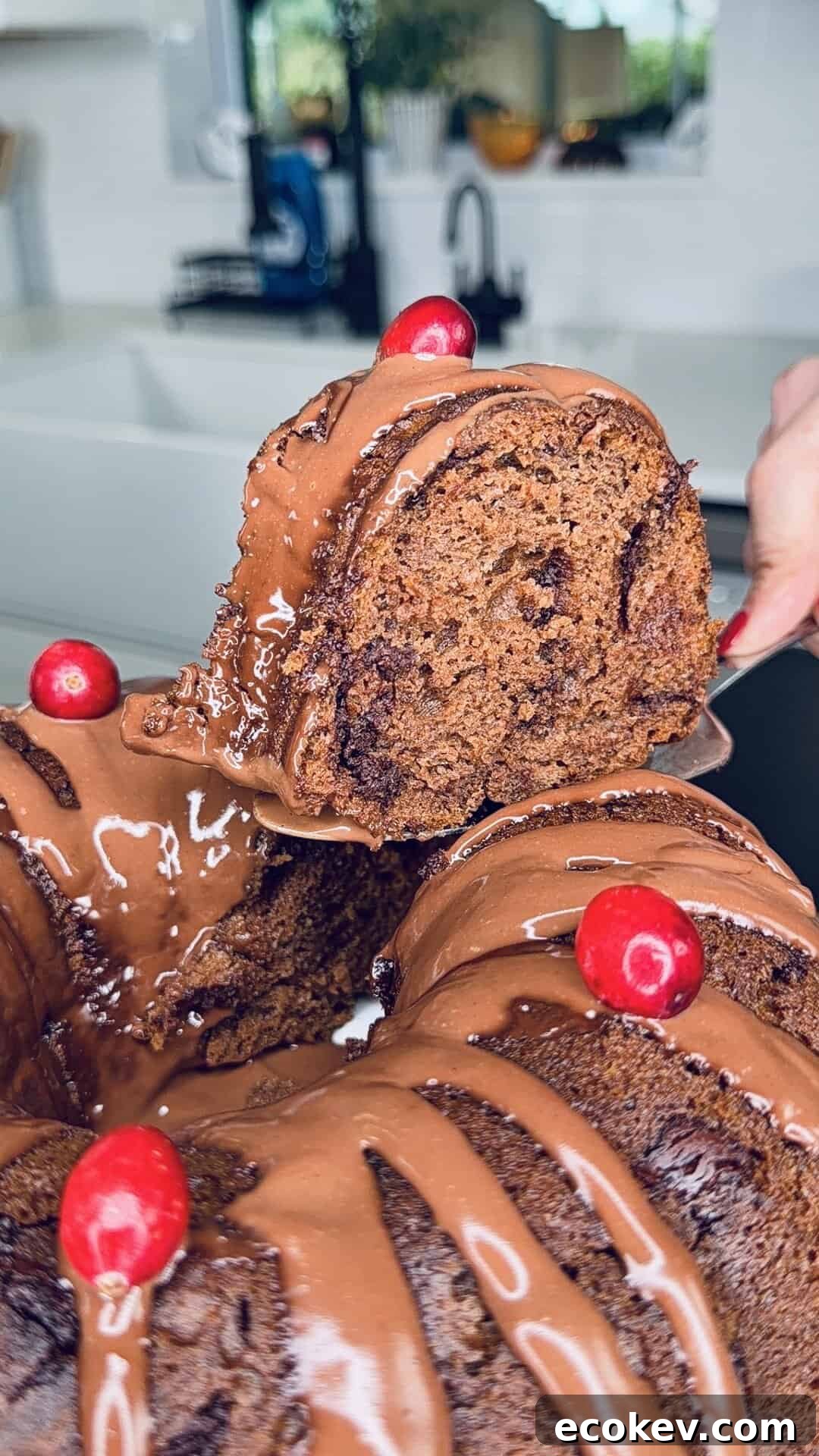 A close-up of a chocolate bundt cake drizzled with chocolate glaze, topped with red cherries. A hand is lifting a slice, revealing the cake’s moist, rich texture. The background shows a modern kitchen.