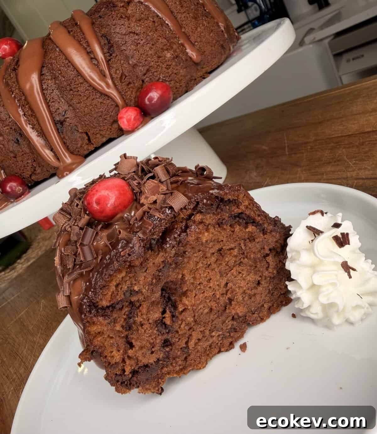 A slice of chocolate bundt cake topped with chocolate shavings and a cherry, served with a dollop of whipped cream on a white plate. The whole cake, decorated similarly, is displayed in the background on a stand.