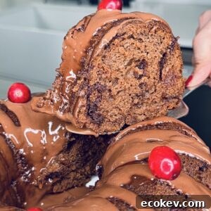 A close-up of a chocolate bundt cake topped with glossy chocolate icing and red berries. A hand is lifting a slice, revealing the moist, rich texture inside. A modern kitchen is blurred in the background.