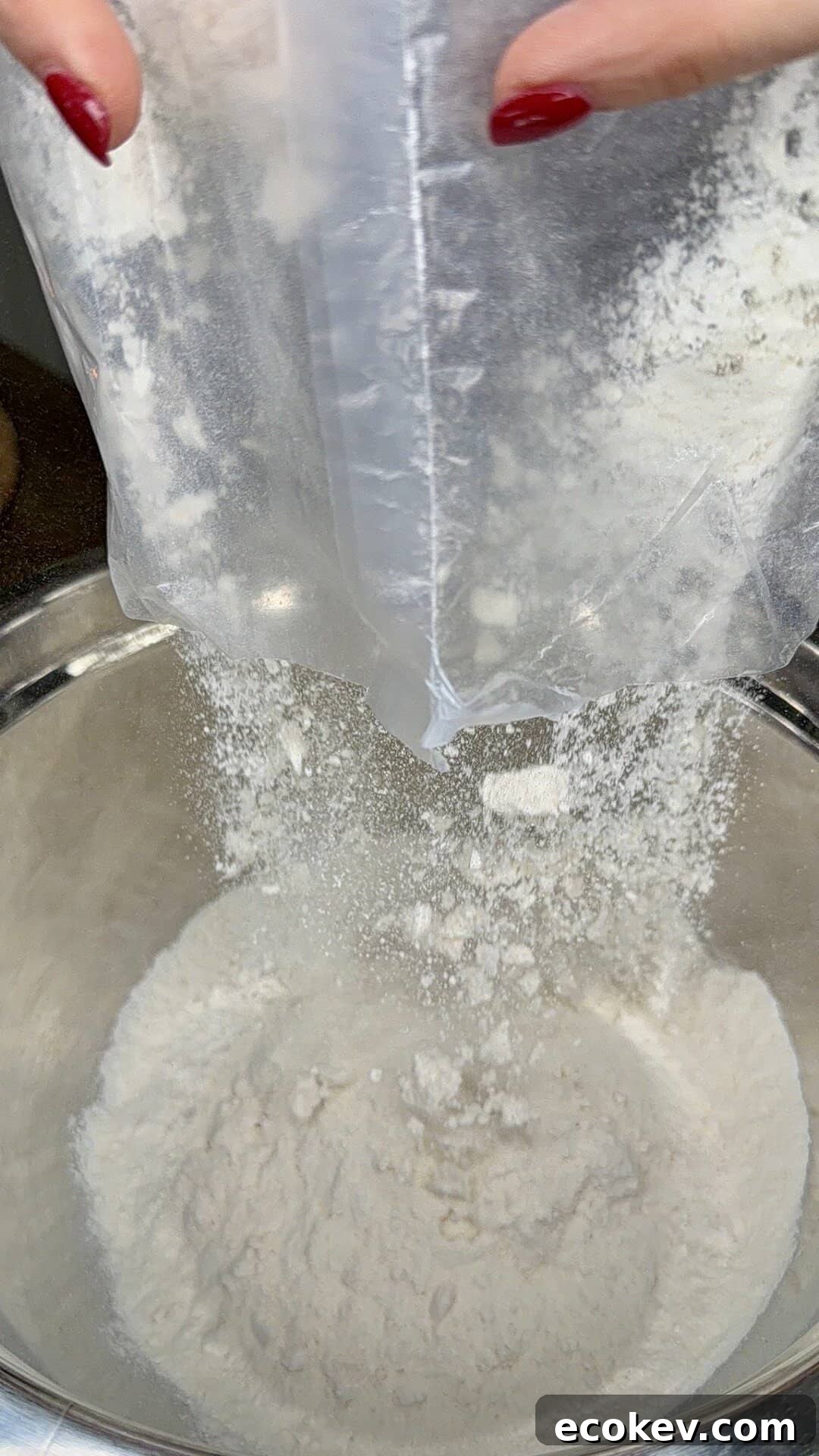 A close-up view of flour being poured from a plastic bag into a metal mixing bowl, with hands holding the bag and red nail polish visible.