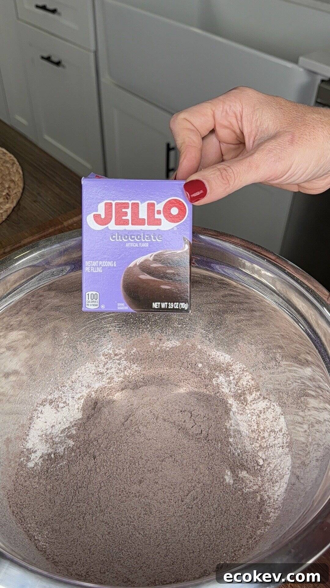 A hand holds a box of Jell-O chocolate instant pudding mix above a large metal bowl containing chocolate powder. Kitchen cabinets and a countertop are visible in the background.