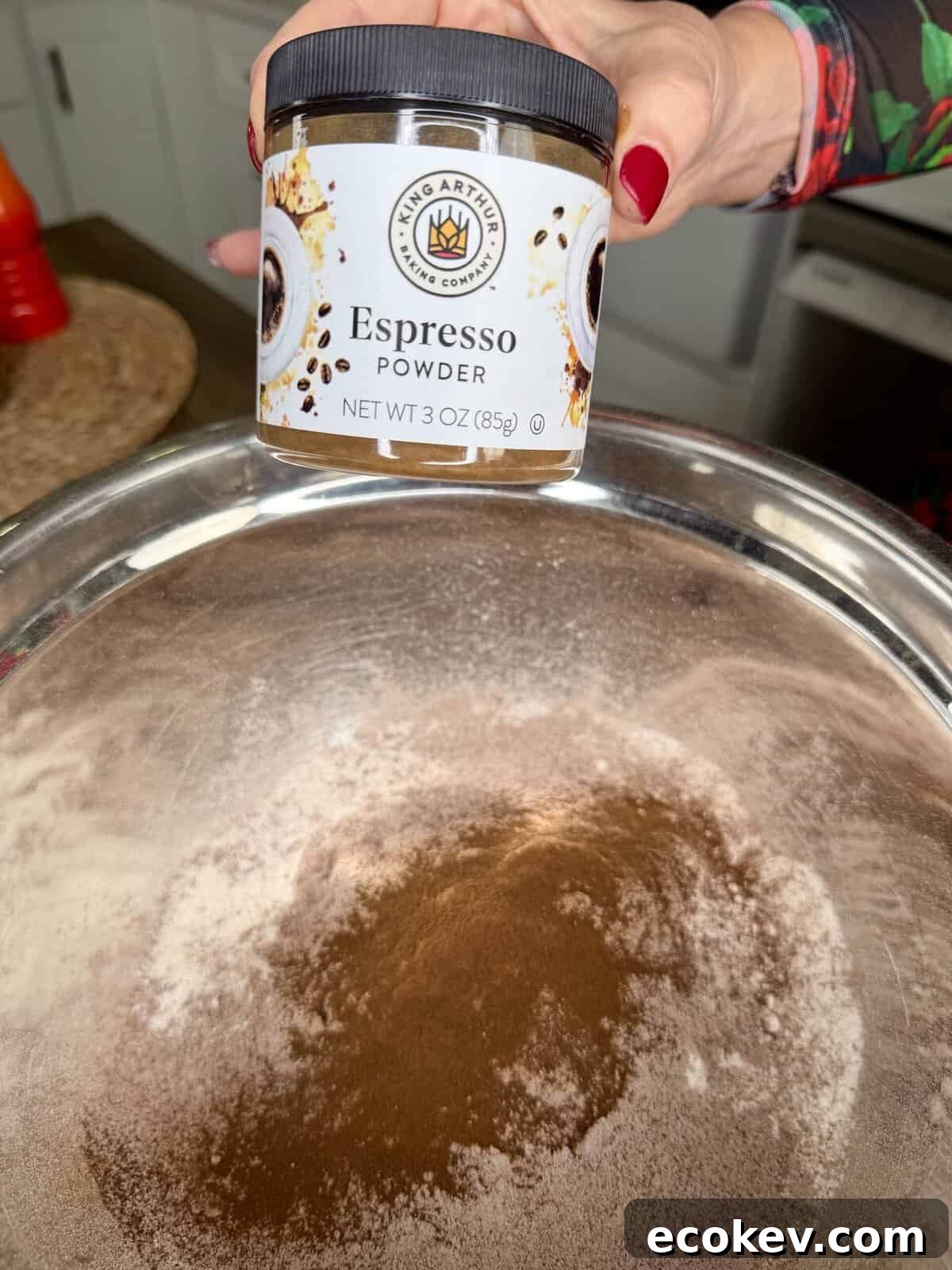 A hand holds a jar of King Arthur Espresso Powder above a large metal bowl filled with flour and cocoa powder, ready for baking.