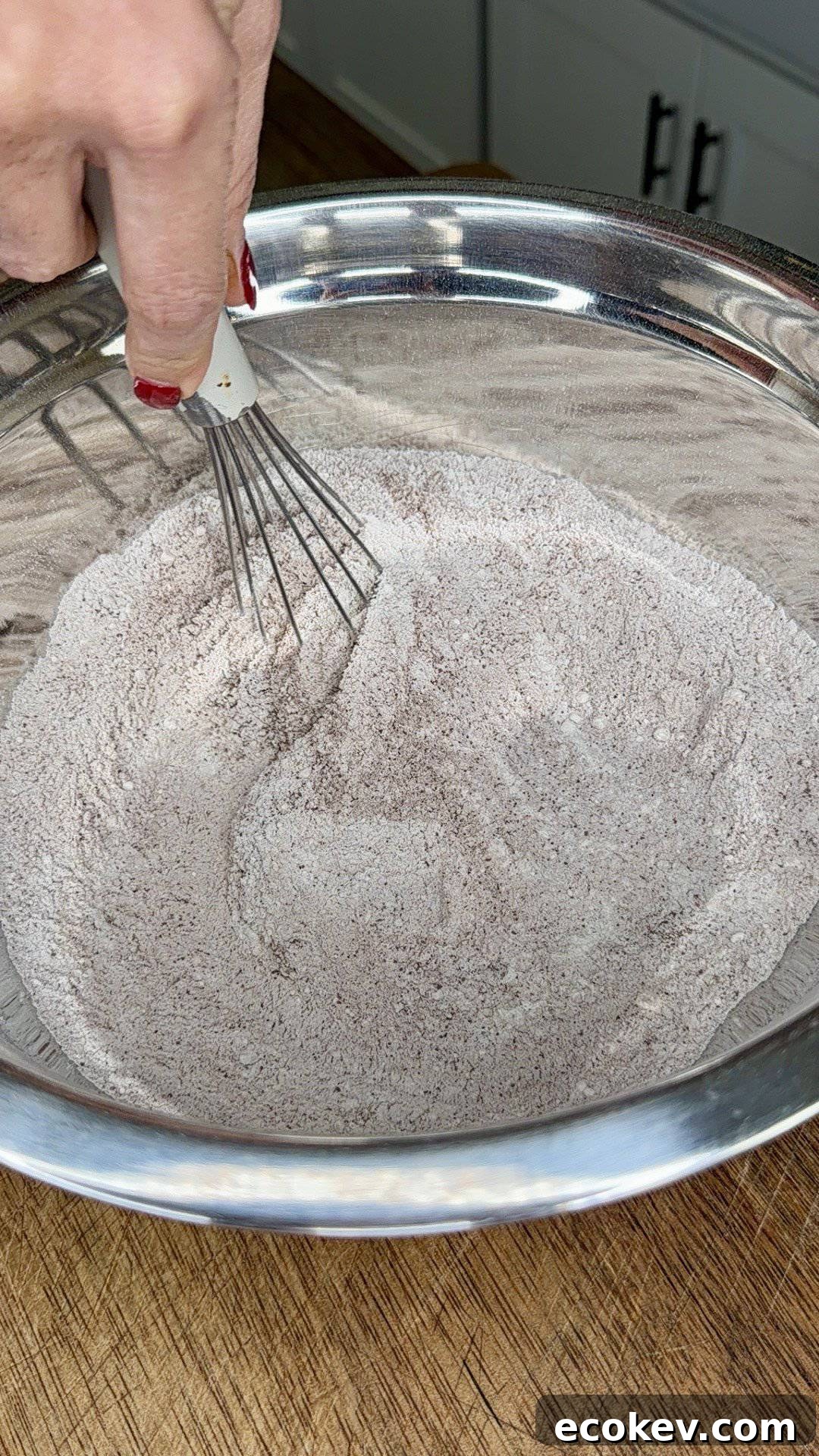 A hand with red nail polish uses a whisk to mix a bowl of dry ingredients, likely flour and cocoa powder, on a wooden countertop.