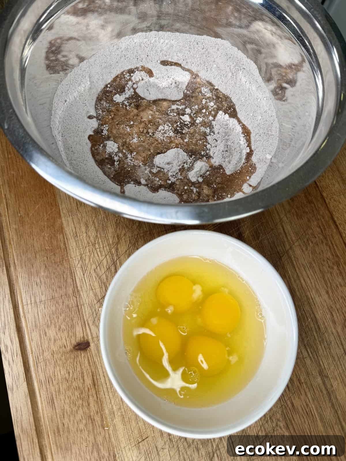 A metal bowl with dry baking ingredients and a mound of brown sugar sits next to a white bowl containing four cracked eggs on a wooden surface.