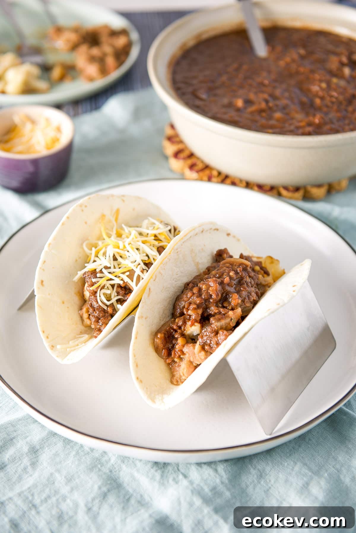 A white plate with a taco holder with flour tortillas and the taco filling in it along with a bowl of lentils in the back