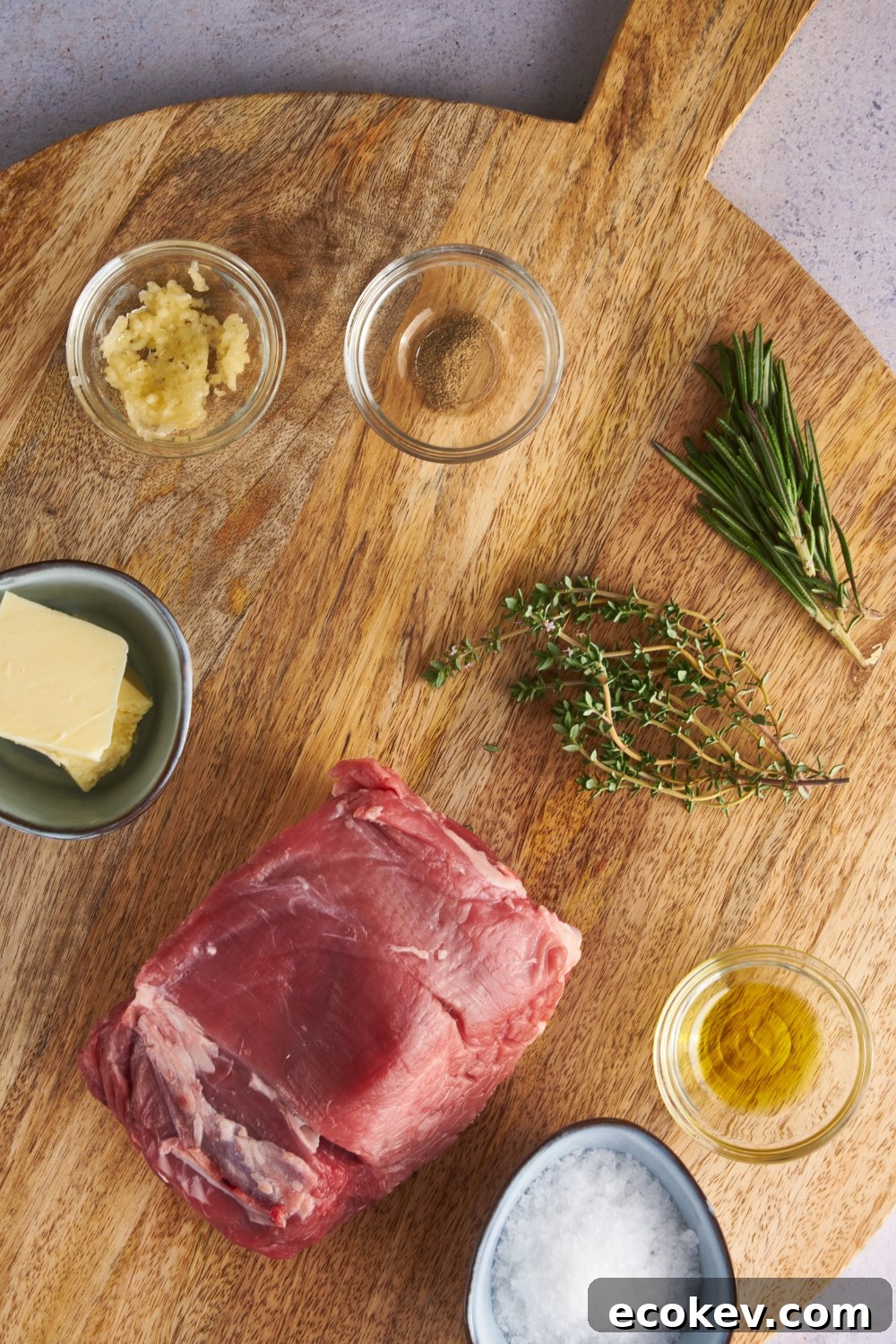 An assortment of ingredients including a raw beef tenderloin, fresh herbs, and bowls of minced garlic, spices, oil, and butter.