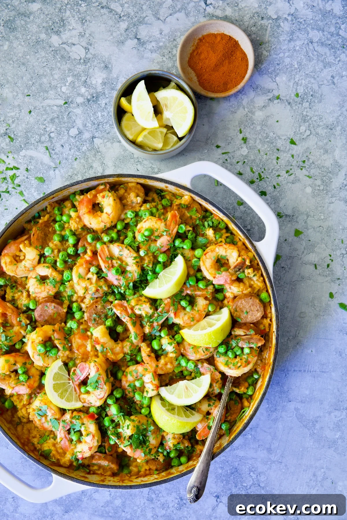 Skillet of Brown Rice Paella with Shrimp and Chorizo with lemon wedges and bowl of paprika.