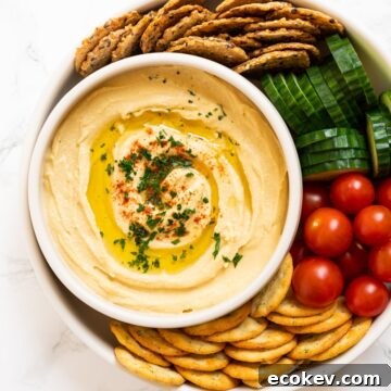 a top down view of a bowl of chickpea hummus surrounded by crackers and veggies.