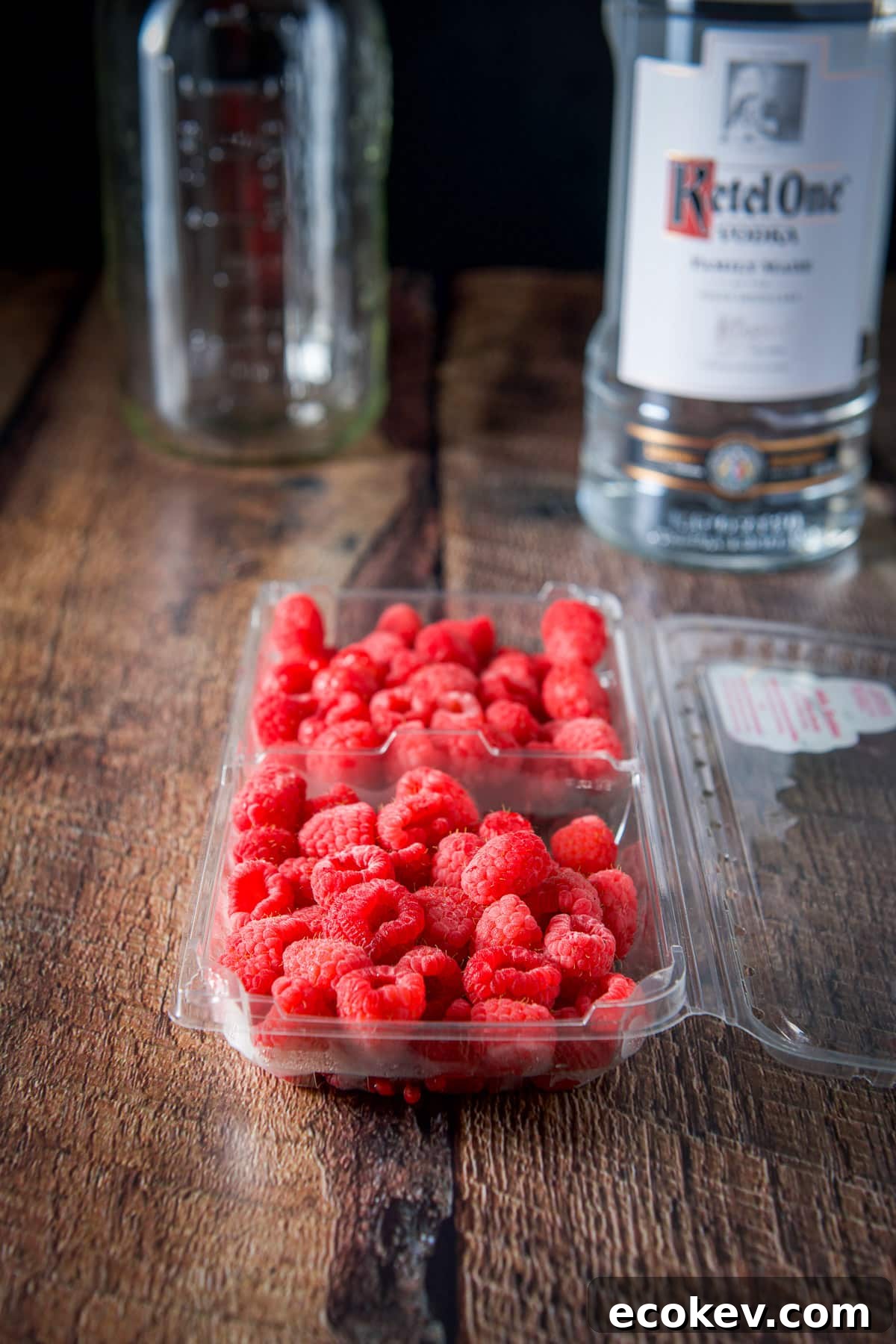 Raspberries in a plastic container, a vodka bottle and jar in the background