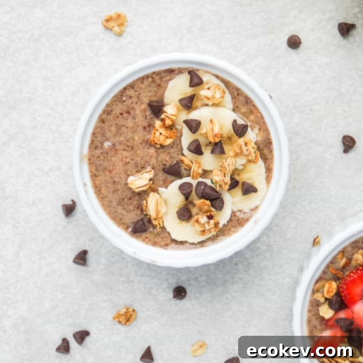 One serving of flaxseed pudding topped with banana slices, granola, and mini chocolate chips in the center, part of a second serving is showing in the bottom right corner, and mini chocolate chips and granola pieces are scattered around the surface.