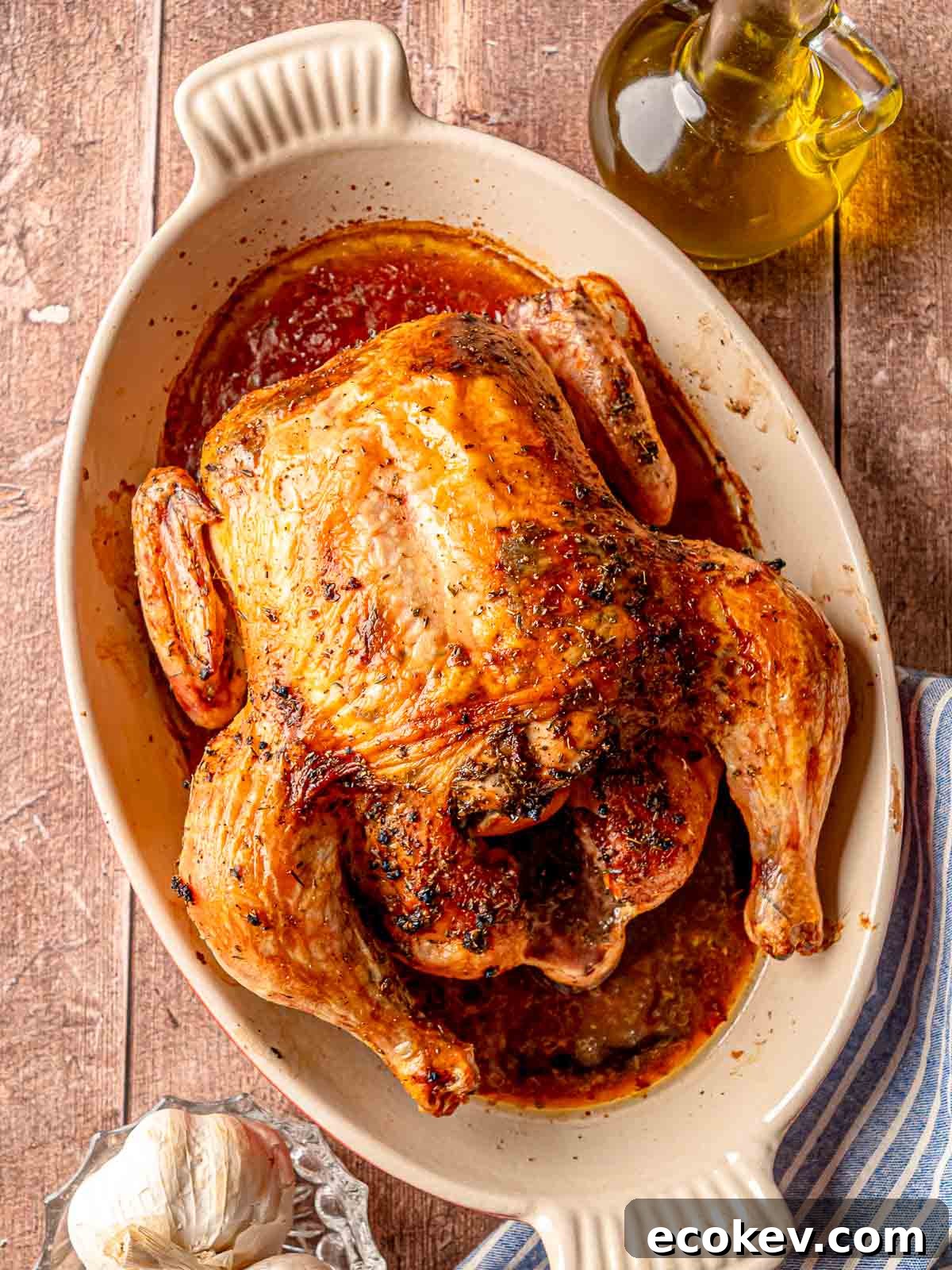 A finished Greek roasted chicken in a baking dish, styled on a wooden surface with a bottle of olive oil, a head of garlic, and a blue striped napkin.