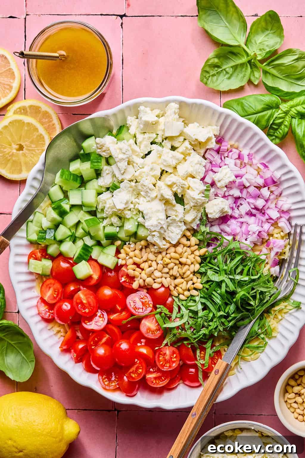 A large white bowl filled with chopped cucumbers, cherry tomatoes, red onion, feta cheese, pine nuts, and shredded basil—this vibrant spread is perfect for a lemon basil orzo salad.