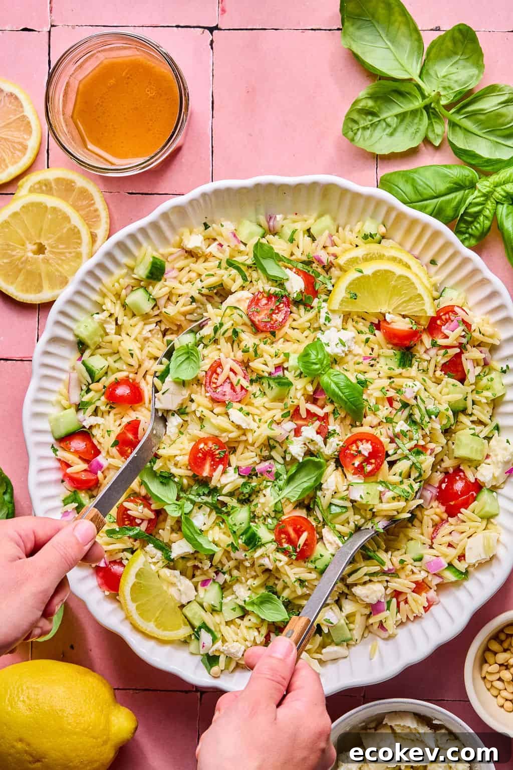 A person tosses a vibrant lemon basil orzo salad with cherry tomatoes, cucumber, red onion, feta, and lemon slices. Nearby, a jar of dressing, lemon halves, basil leaves, and pine nuts sit on a pink tiled surface.