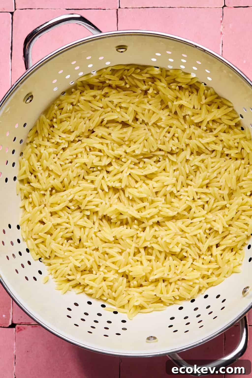 Cooked orzo pasta, perfect for a lemon basil orzo salad, is draining in a metal colander set on a pink tiled surface.