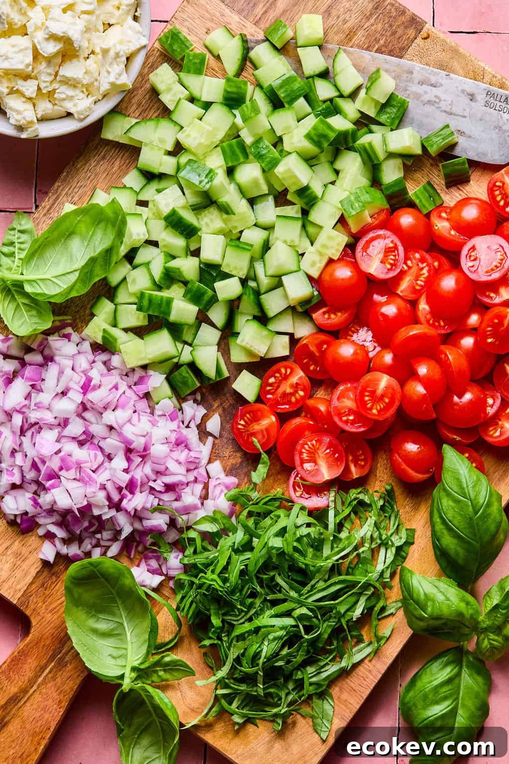A wooden cutting board with chopped cucumber, halved cherry tomatoes, diced red onion, shredded basil, and whole basil leaves.