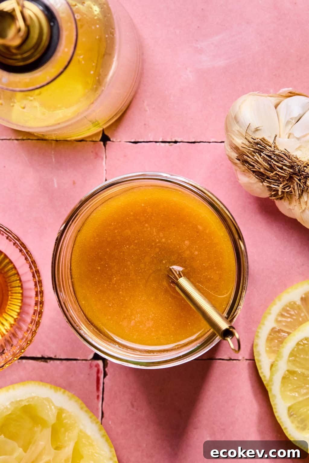 A glass jar filled with honey mustard dressing sits with a spoon inside, surrounded by a lemon half, garlic head, oil bottle, and small bowl on a pink tiled surface.