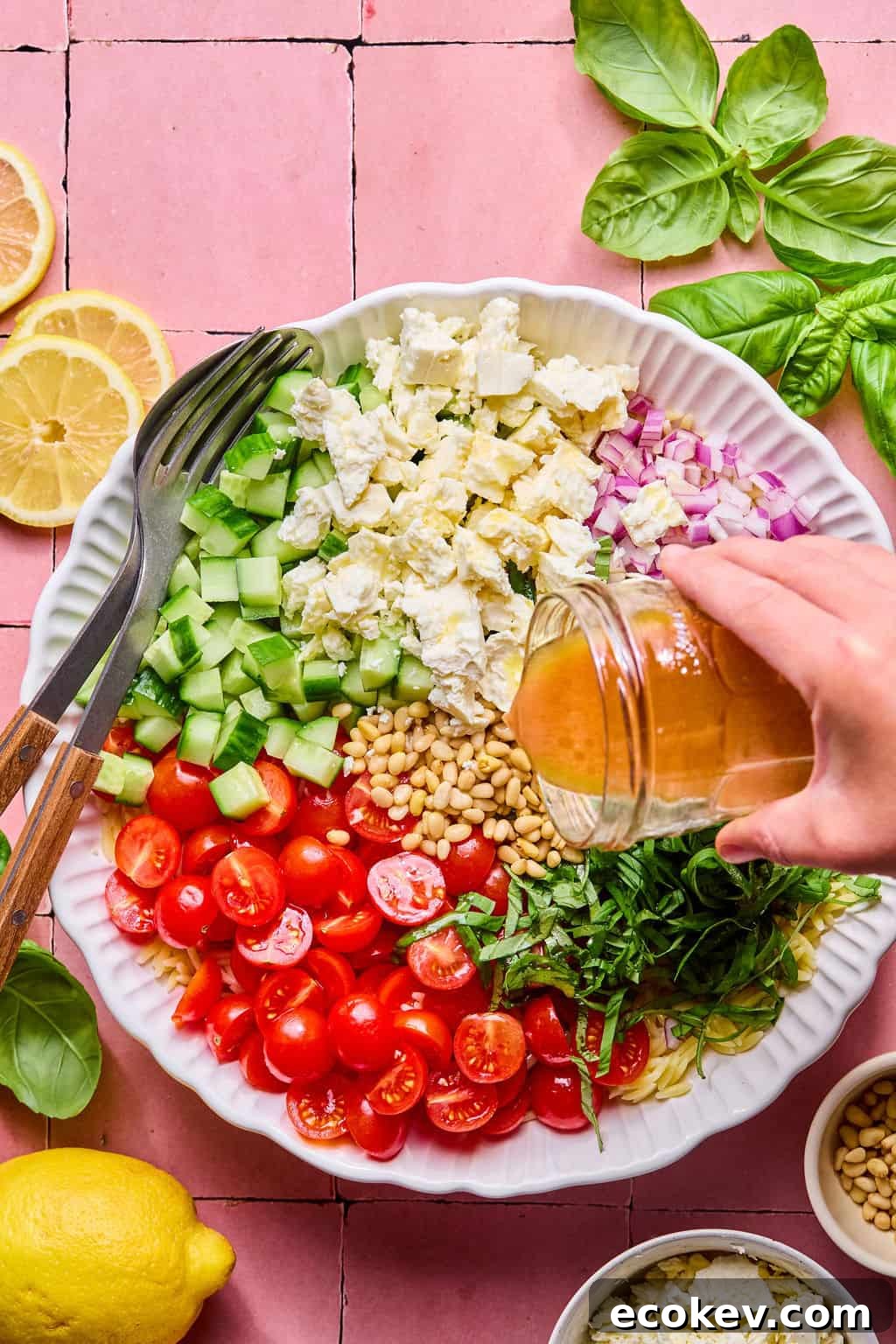 A hand pours dressing from a jar over a vibrant lemon basil orzo salad with cherry tomatoes, cucumber, red onion, feta, pine nuts, and basil in a white bowl.