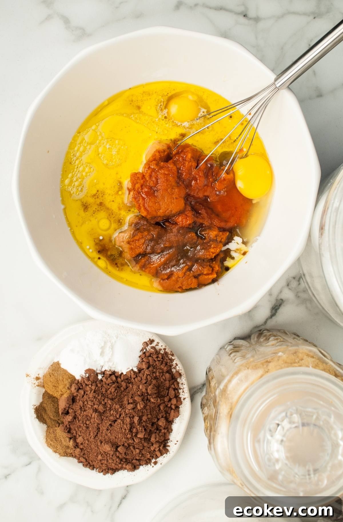 A bowl with eggs, pumpkin puree, and other ingredients being whisked, next to dishes of cocoa and spices—perfect for making a festive pumpkin bundt cake or Halloween pumpkin cakes.