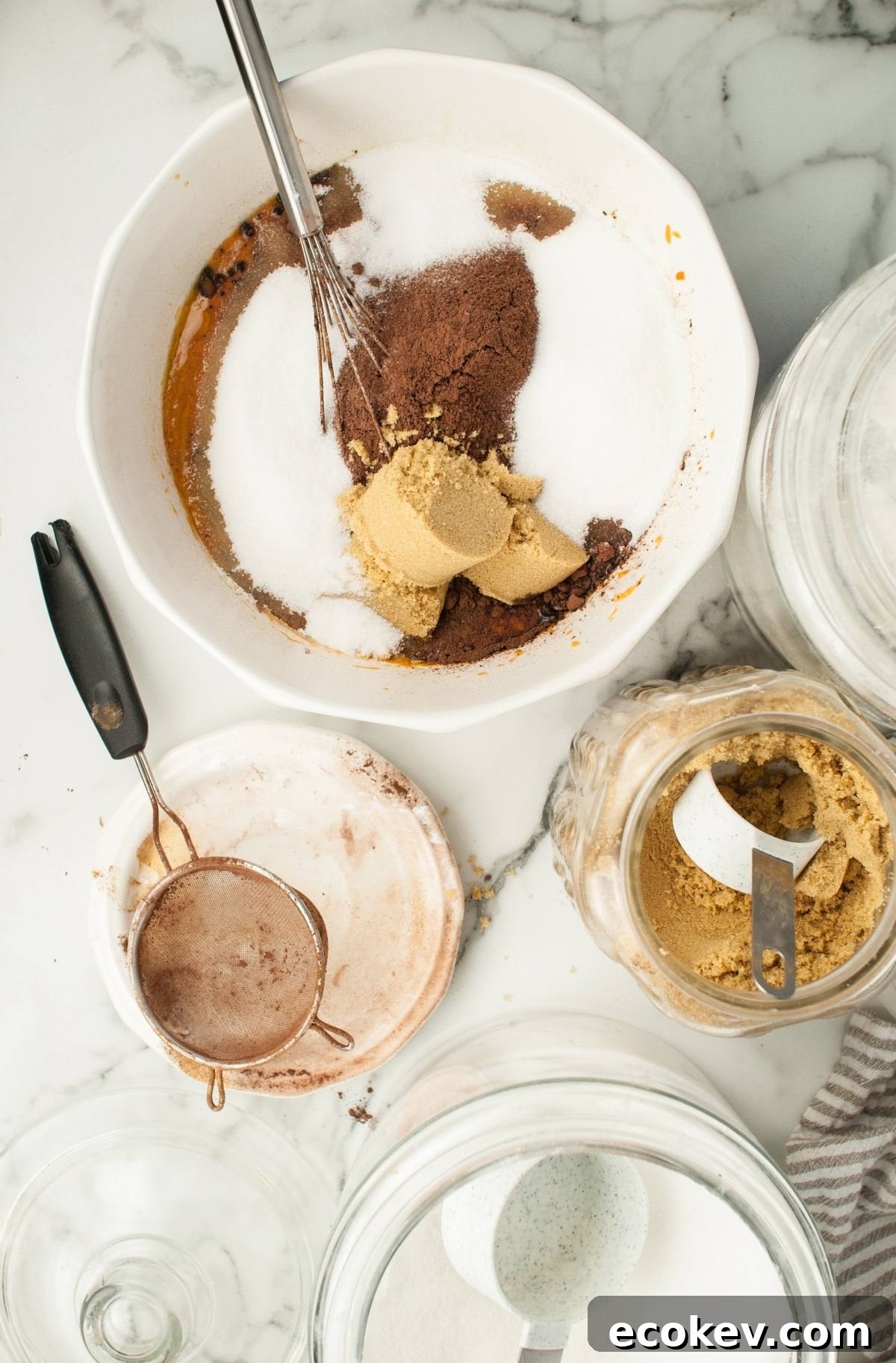 Baking ingredients in a bowl with a whisk; brown sugar, white sugar, and cocoa powder on a marble surface—perfect for making Pumpkin Shaped Mini Bundt Cakes as a festive Thanksgiving or Halloween dessert.