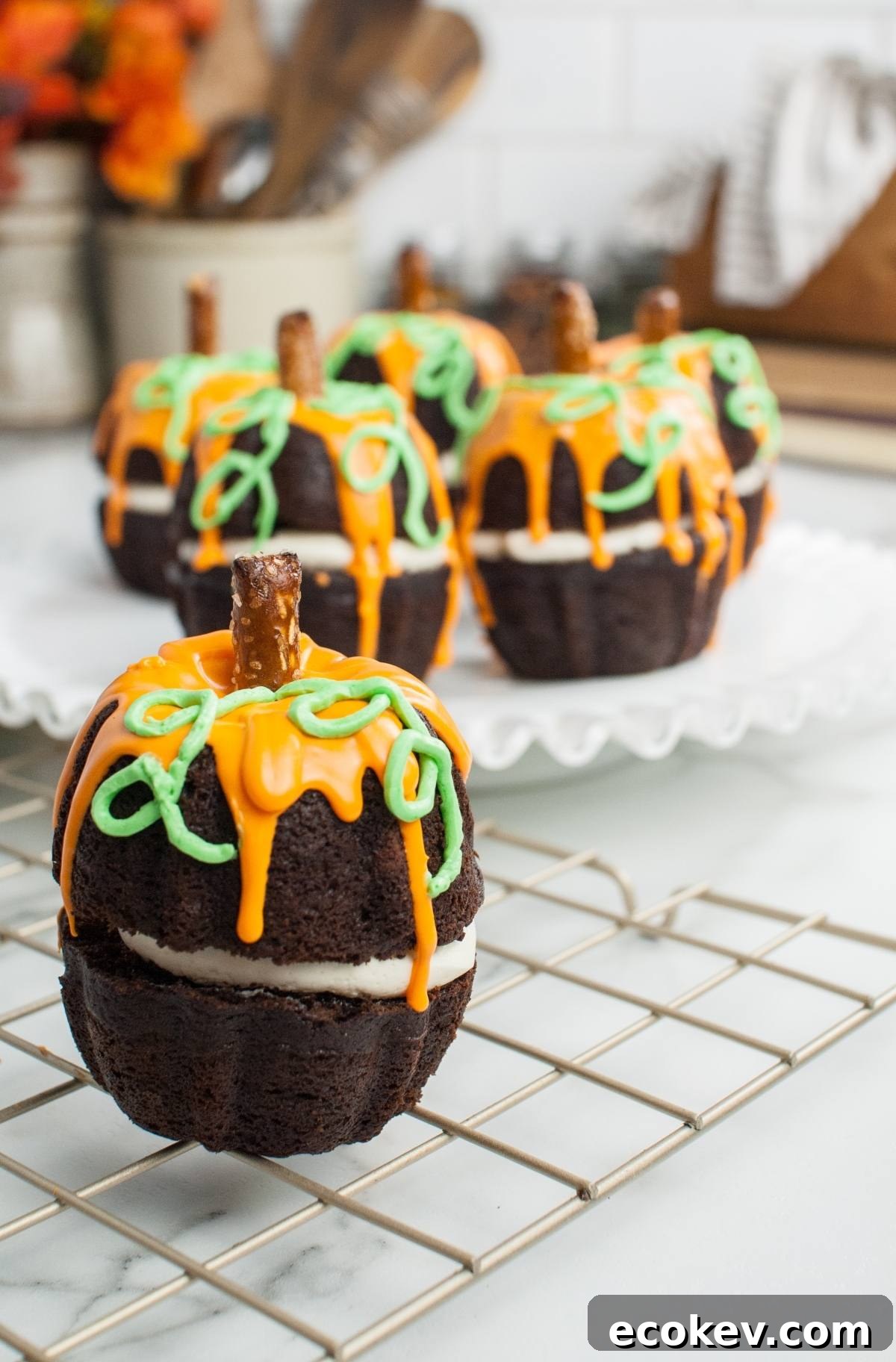 Pumpkin Shaped Mini Bundt Cakes with orange icing and pretzel stems rest on a cooling rack and serving plate—perfect as a festive Thanksgiving or Halloween dessert.