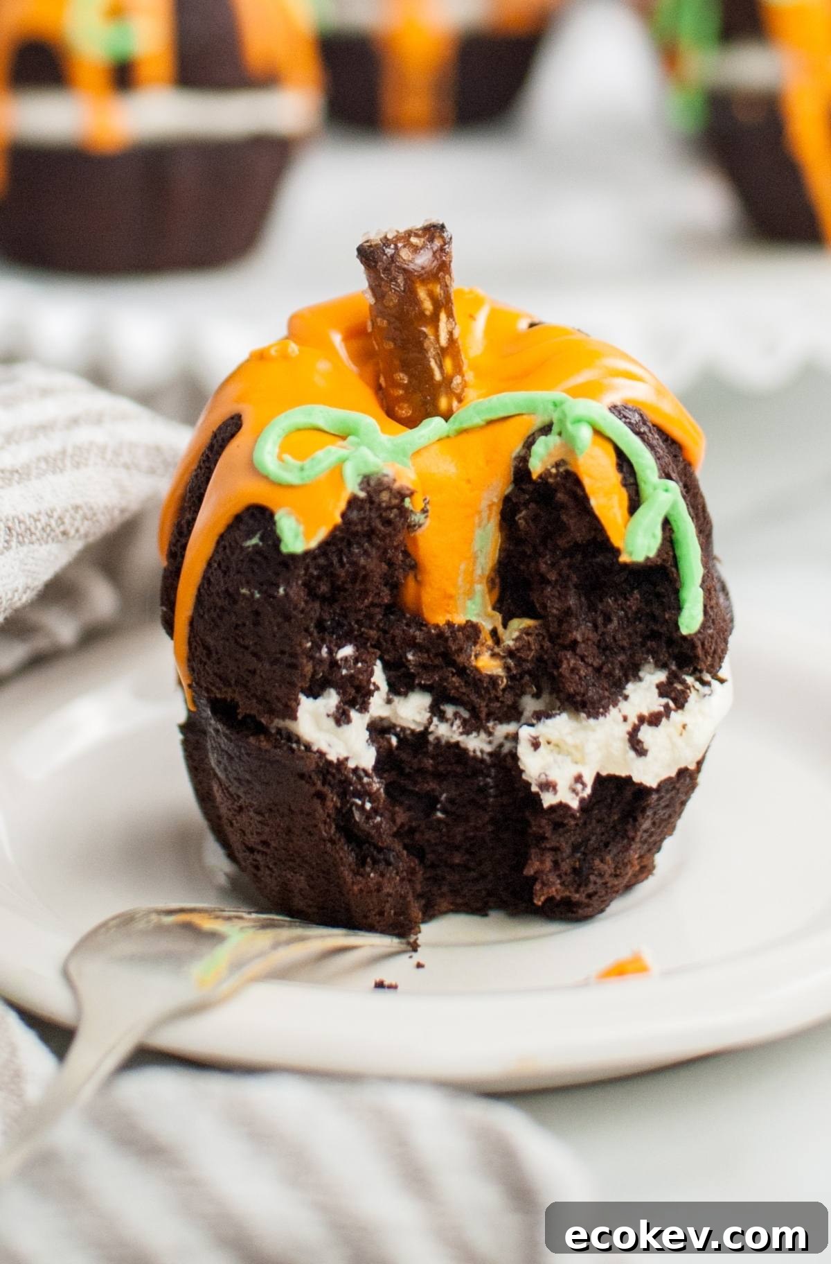 Chocolate cupcake shaped like a pumpkin with orange icing, creamy filling, and a pretzel stem, partially eaten—a playful twist on Halloween pumpkin cakes, perfect as a Thanksgiving or Halloween dessert.