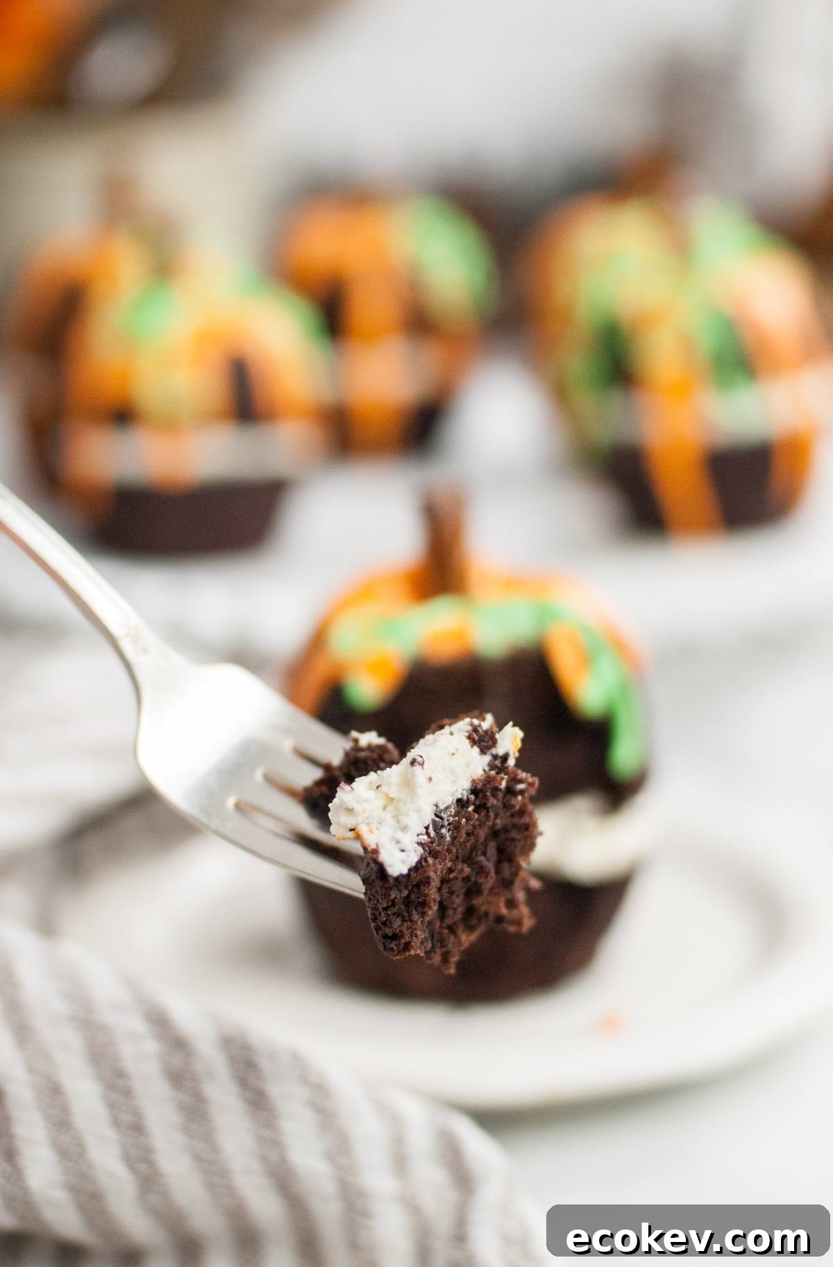 A fork holds a bite of chocolate cupcake with cream filling; in the background, decorated cupcakes and a pumpkin bundt cake set the scene.