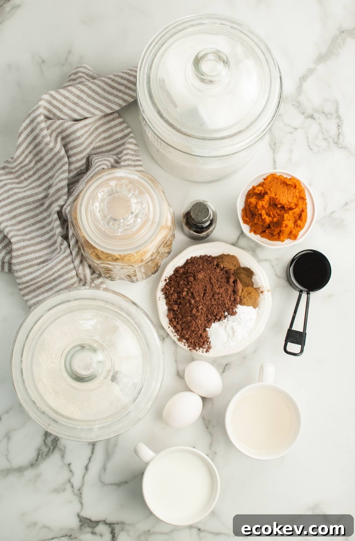 Baking ingredients on marble for Pumpkin Shaped Mini Bundt Cakes: eggs, cocoa, pumpkin puree, sugar, flour jars, vanilla, milk, and measuring cups—perfect for a festive Thanksgiving or Halloween dessert.