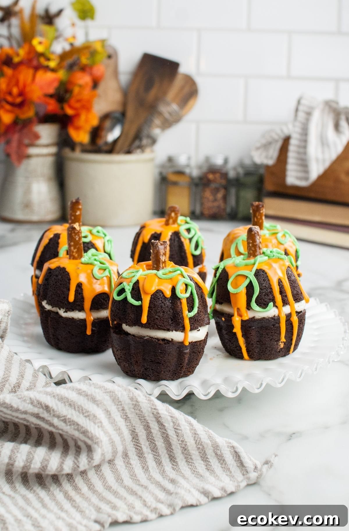 Chocolate cupcakes decorated like pumpkins with orange and green icing, topped with pretzel sticks, make a festive Thanksgiving or Halloween dessert—perfect for fans of pumpkin shaped mini bundt cakes. Displayed on a white cake stand.
