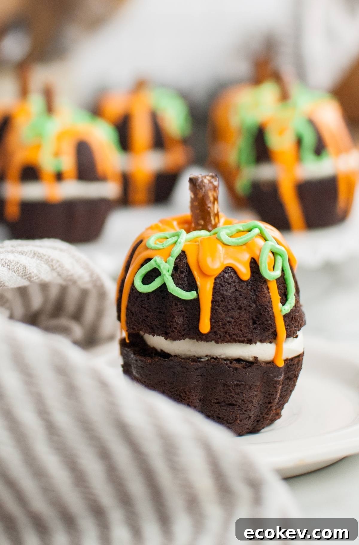 Chocolate cake shaped like a pumpkin, with orange and green icing and a pretzel stem, makes a festive Halloween pumpkin cake that’s perfect as a Thanksgiving or Halloween dessert. Served on a white plate.