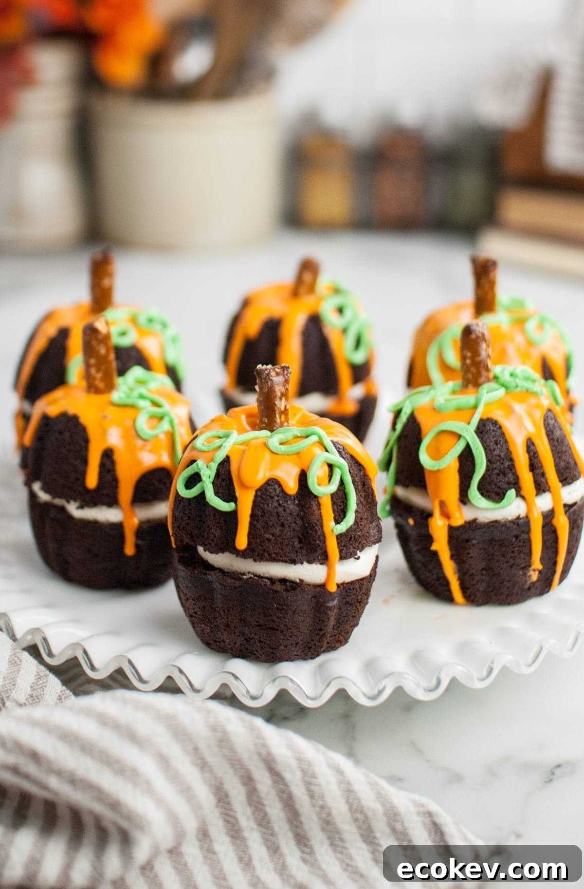 Pumpkin Shaped Mini Bundt Cakes in rich chocolate, topped with orange and green icing and pretzel stems, make a delightful Thanksgiving or Halloween dessert on a white plate.