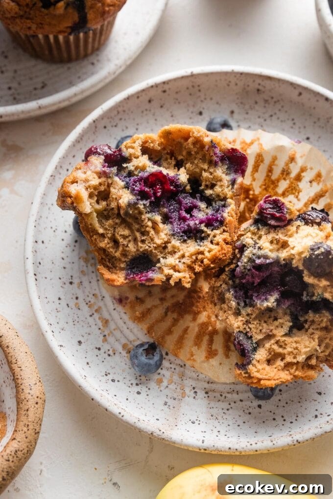 Small white speckled ceramic plate with a blueberry banana muffin pulled apart to show the moist interior and crunchy top.