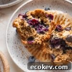 Small white speckled ceramic plate with a blueberry banana muffin pulled apart to show the moist interior and crunchy top.
