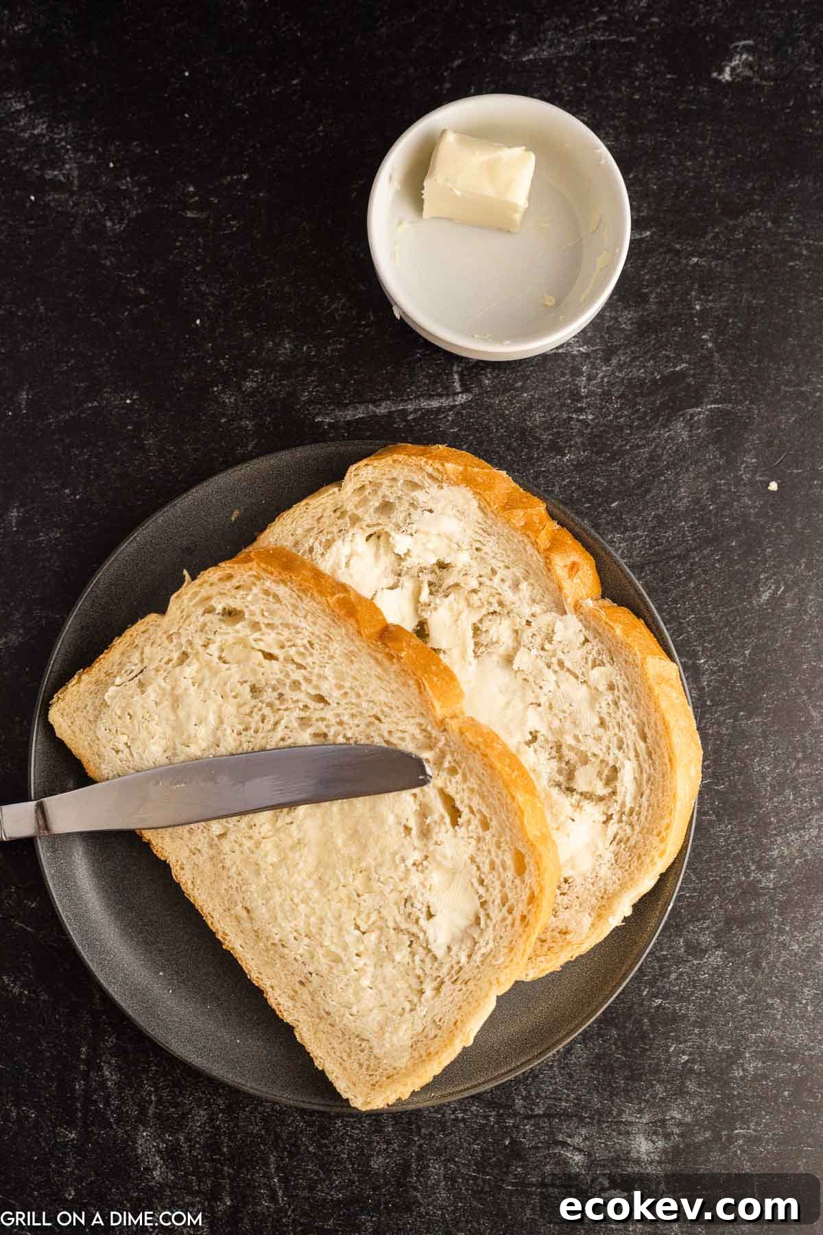 Two slice of bread with butter spread on them on a black plate with a bowl of butter on the side