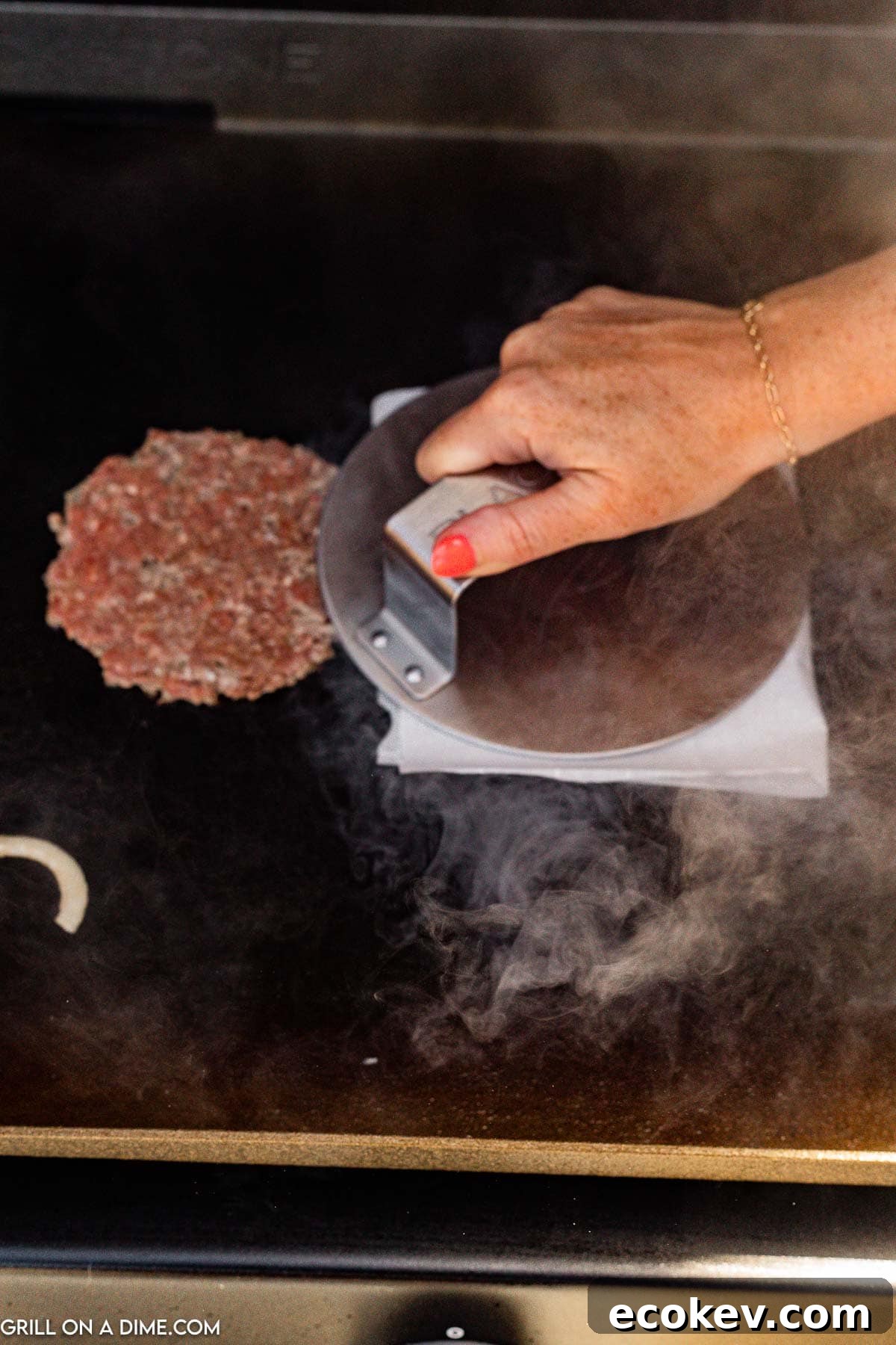 Ground beef patties on the griddle with a burger press on parchment paper pressing on the ground beef patty on the griddle
