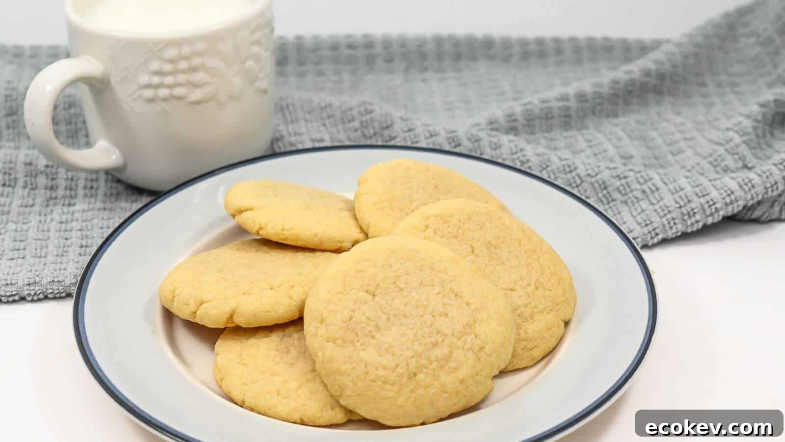 A plate of sugar cookies with a mug of milk and a gray cloth in the background.