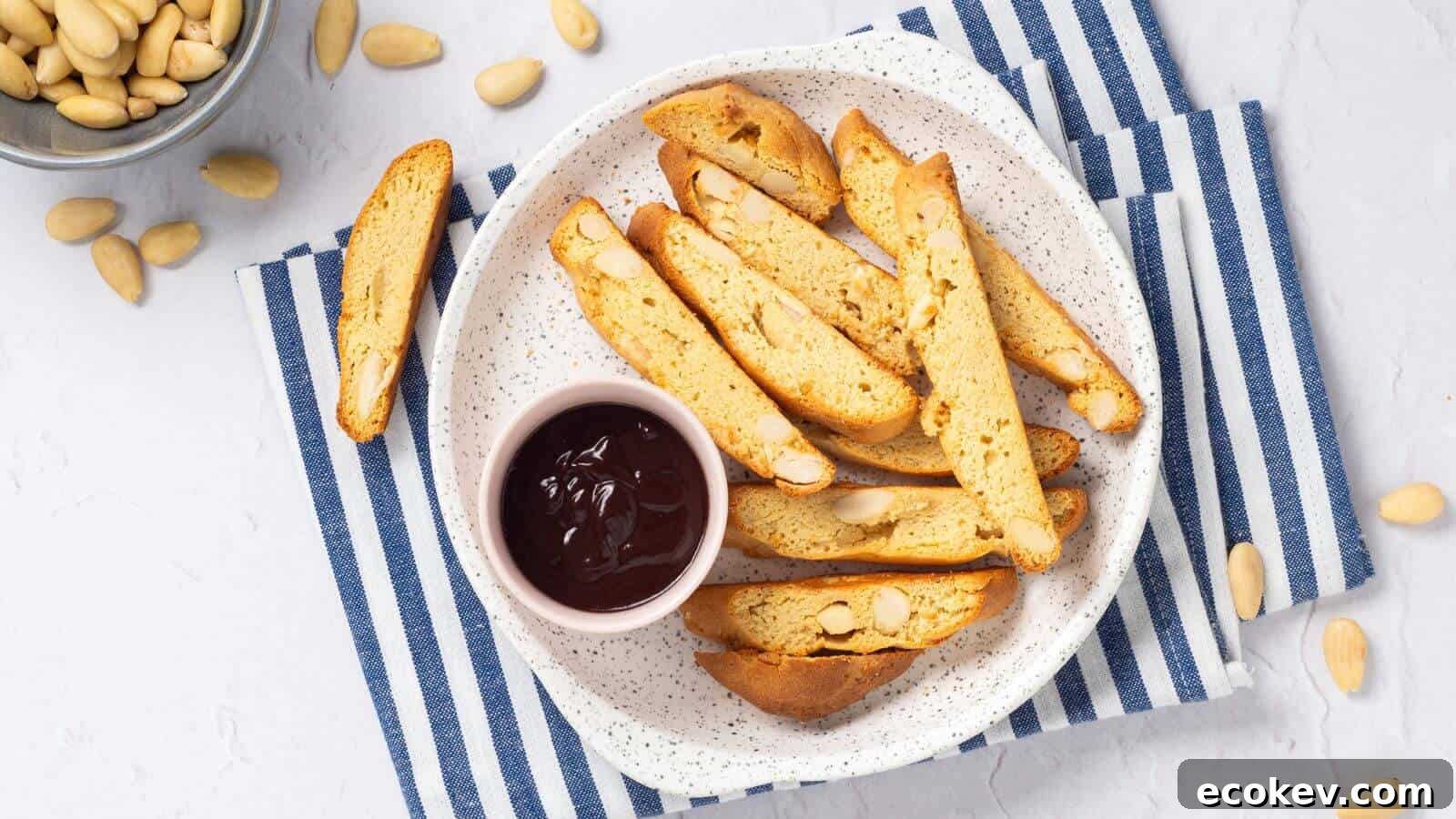 A plate of almond biscotti with a small bowl of chocolate sauce on a blue and white striped cloth. Almonds are scattered around.