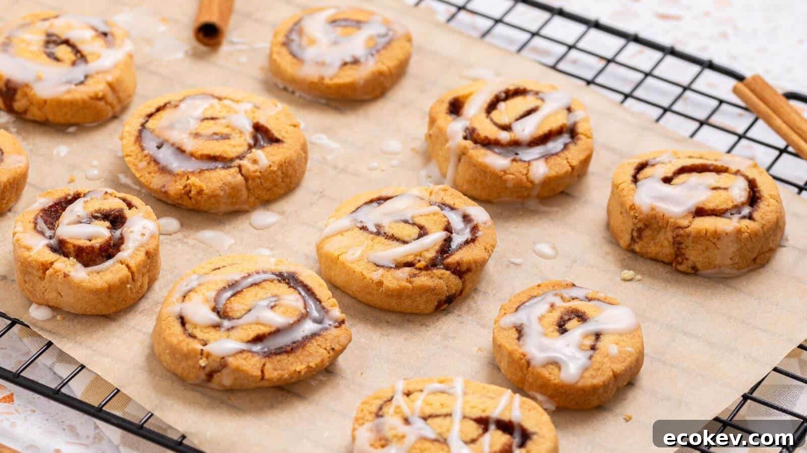 Cinnamon roll cookies with icing are placed on a sheet of parchment paper on a cooling rack. Two cinnamon sticks are visible beside them.