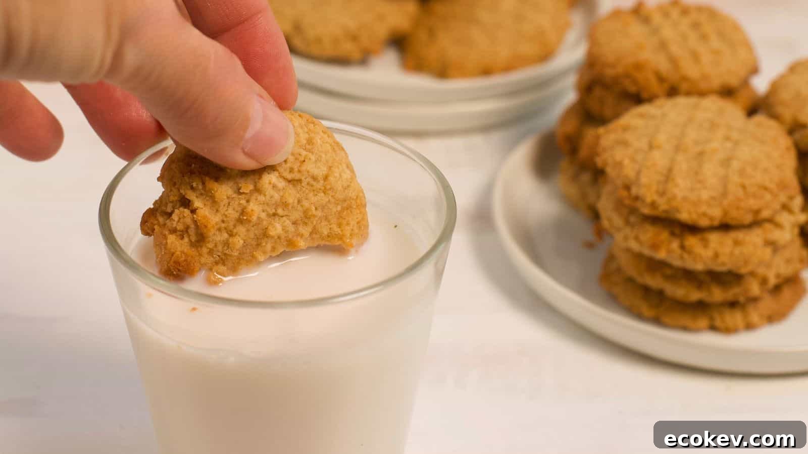 A person dipping a peanut butter cookie into a glass of milk, with more stacked cookies on a plate in the background.