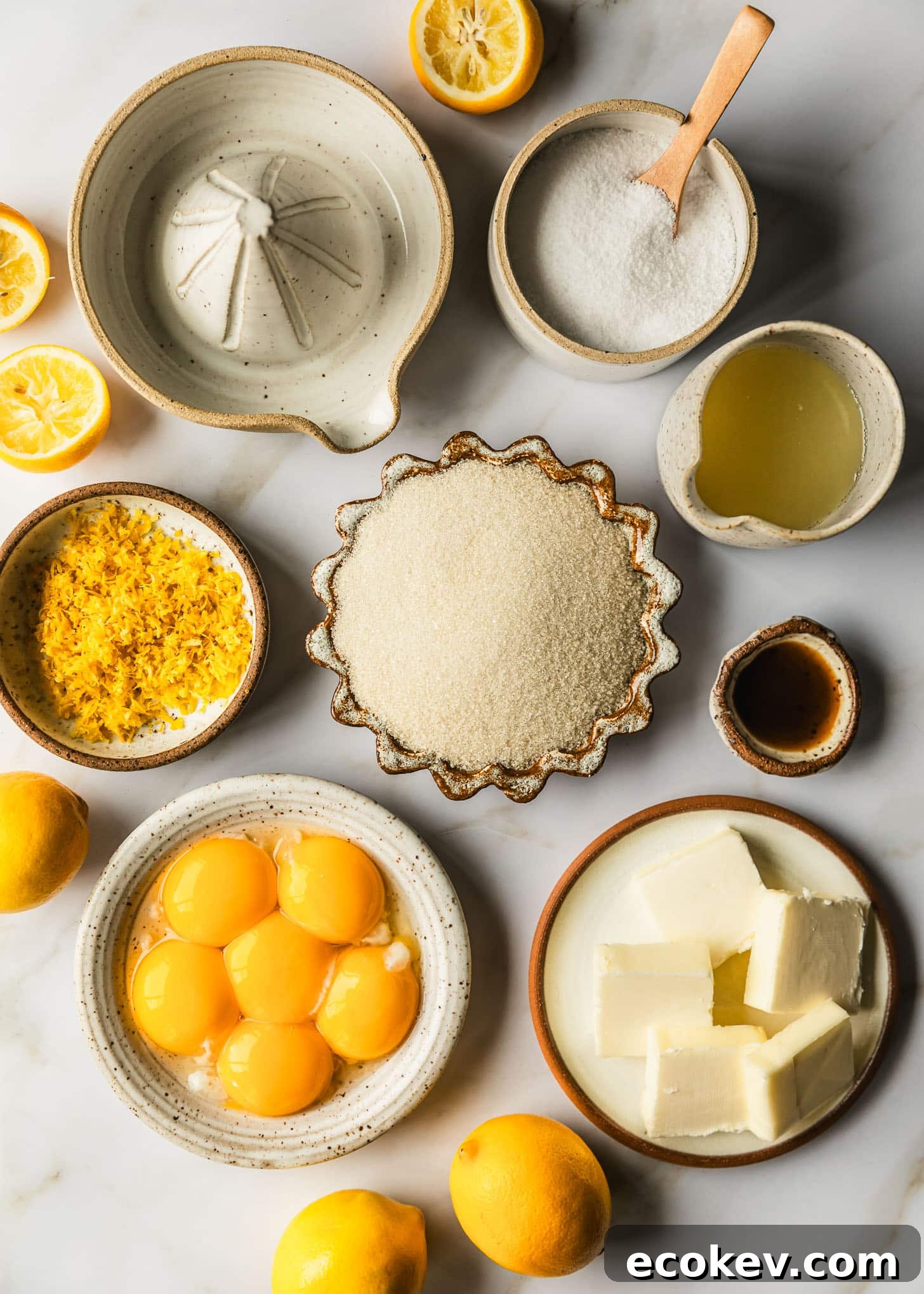 White and brown bowls of salt, sugar, lemon zest, lemon juice, vanilla, butter, and egg yolks on a marble counter next to lemons.